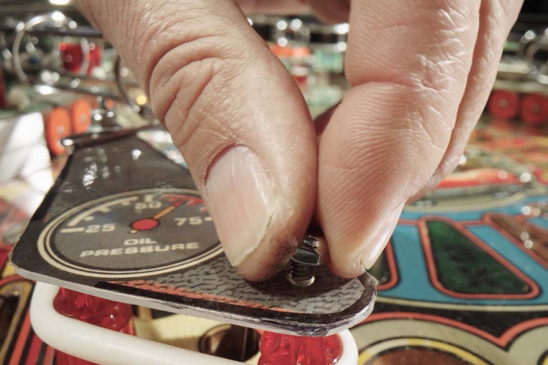 Eine Person schraubt eine Abdeckung mit einer Öldruckanzeige an einem Flipperautomaten fest.
