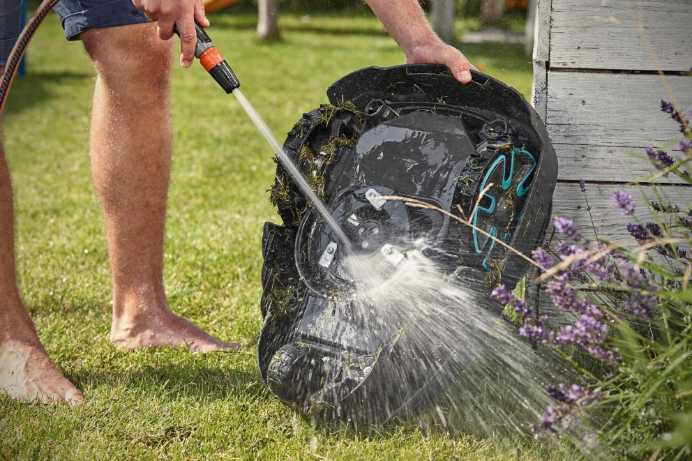 Eine Person reinigt einen Rasenmähroboter mit einem Wasserschlauch im Garten.