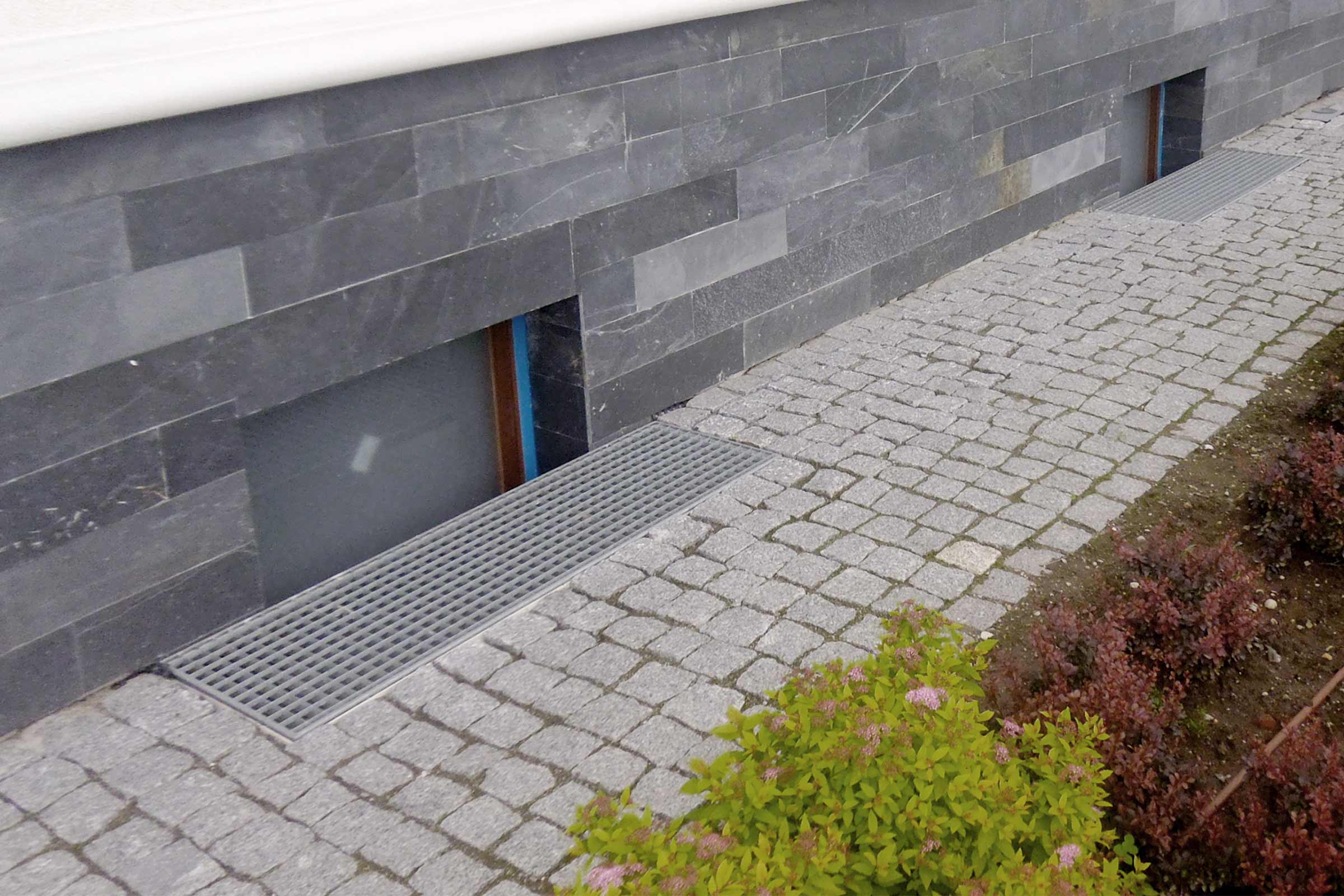 Außenansicht eines Hauses mit Steinfassade, gepflastertem Weg und Entwässerungsrinne vor den Fenstern