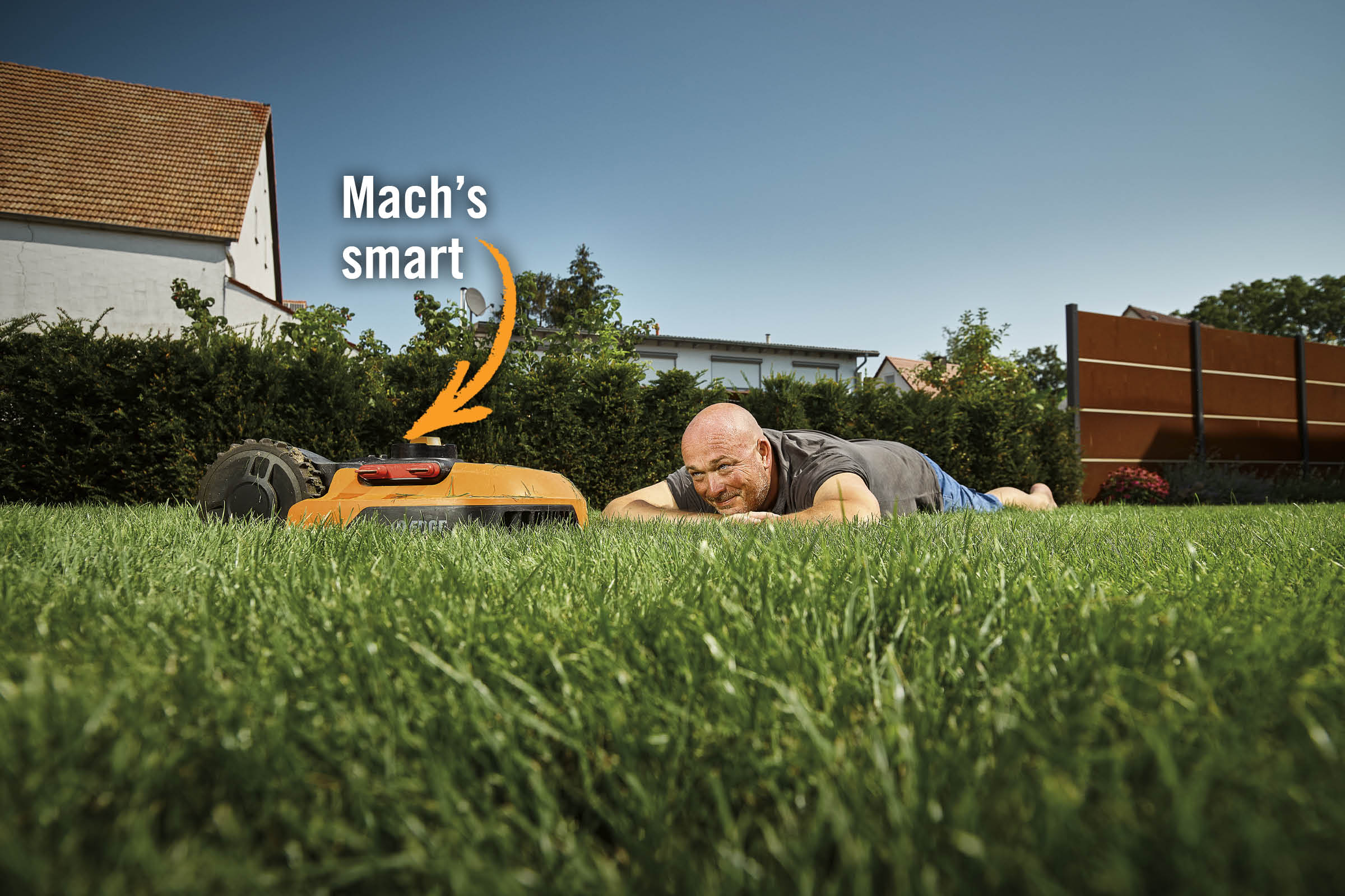 Ein Mann liegt im Gras neben einem Rasenmähroboter auf einer Rasenfläche.