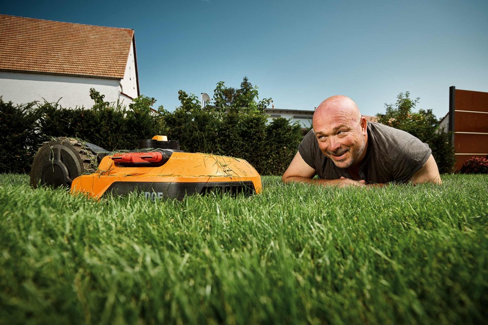 Ein Mann liegt im Gras neben einem Mähroboter im Garten.