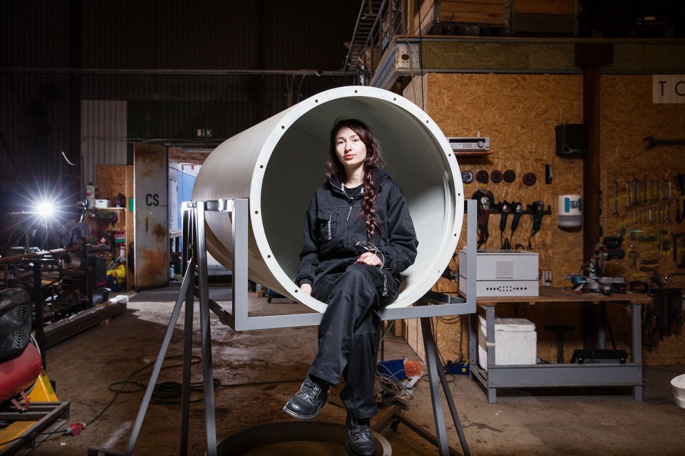 Eine Frau sitzt in einem großen Rohr auf einem Metallgestell in einer Werkstatt.