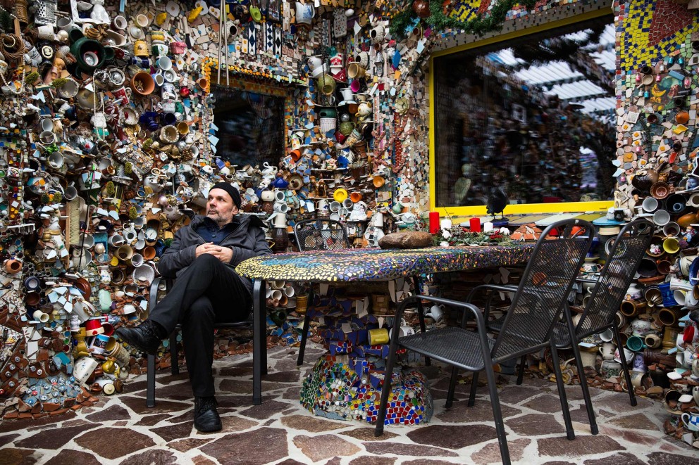 Steffen Modrach sitzt im Hinterhof seines selbst erbauten Hundertwasser Schlosses Lilllliput an einem Tisch, der ebenfalls mit Mosaiksteinen verziert ist. Steffen Modrach sitzt im Hinterhof seines selbst erbauten Hundertwasser Schlosses Lilllliput an einem Tisch, der ebenfalls mit Mosaiksteinen verziert ist.