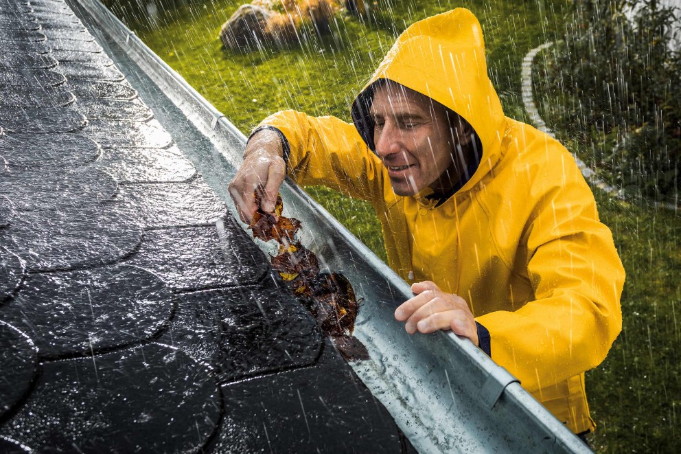 Mann in Regenkleidung reinigt Dachrinne von Laub