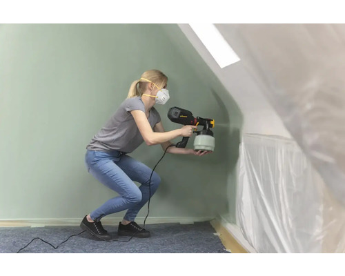 Eine Frau verwendet ein Farbspritzgerät in einem Raum, der mit Folie abgedeckt ist.