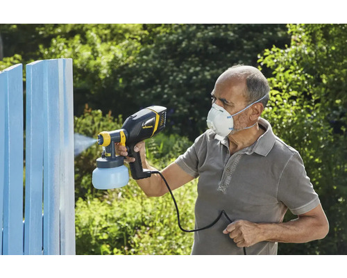 Eine Person verwendet ein Farbsprühgerät, um einen Holzzaun zu streichen. Die Person trägt eine Schutzmaske.