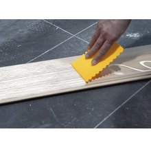 Eine Person verteilt Klebstoff mit einem Spachtel auf einem Holzbrett.