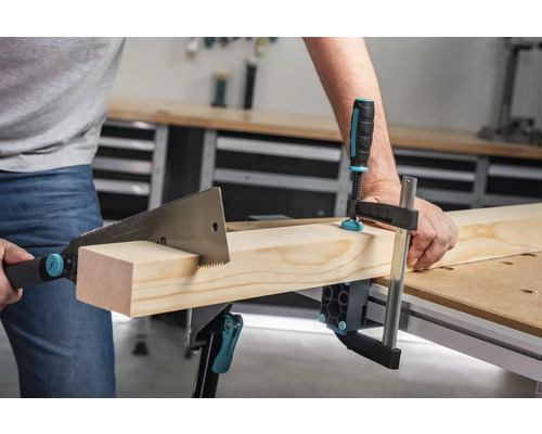 Eine Person sägt ein Holzstück, das mit einer Schraubzwinge an einem Werktisch befestigt ist.