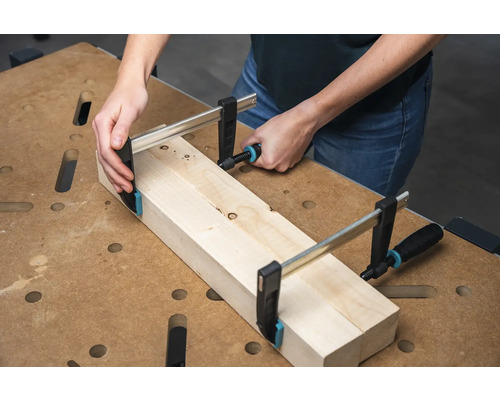 Eine Person befestigt zwei Holzstücke mit einer Schraubzwinge auf einer Werkbank.