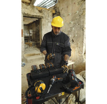 Handwerker mit gelbem Schutzhelm an einem Tisch mit Akkus, Ladegeräten und Werkzeugen in einem Gebäude.