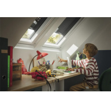Kind schaltet an einem Schreibtisch in einem Dachzimmer das Licht ein. Zu sehen sind außerdem Dachfenster mit Verdunkelungsrollos.
