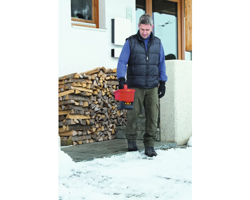 Mann trägt einen Streuwagen der Marke Wolf Garten vor einem Haus mit Holzstapel