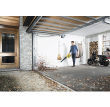 Mann benutzt einen Laubbläser in einer Garage, um Laub zu entfernen. Im Hintergrund sind ein Motorroller und Holzstapel zu sehen.