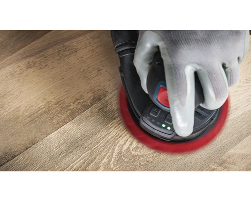 Eine Hand mit Schutzhandschuh bedient einen Exzenterschleifer auf einer Holzoberfläche.