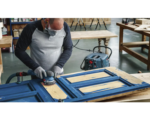 Eine Person schleift mit einem Exzenterschleifer eine blaue Holztür, unterstützt von einem Staubsauger.