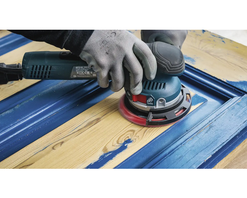 Eine Person verwendet einen Exzenterschleifer auf einem Holzrahmen, um alte Farbschichten zu entfernen.