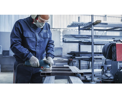 Ein Mann schleift Metall mit einem Schleifgerät, geschützt mit Schutzkleidung in einer Werkstatt.