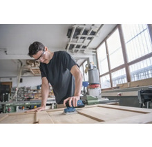 Ein Mann schleift eine Holztür mit einem elektrischen Schleifer in einer Werkstatt.