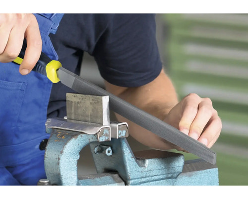 Eine Werkstatt-Szene zeigt einen Mann, der eine Metallfeile mit Griff benutzt, um ein Metallstück in einem Schraubstock zu bearbeiten.