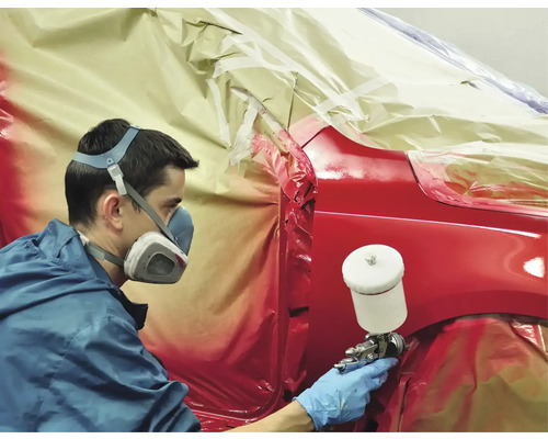 Ein Mann lackiert mit Atemschutzmaske ein rotes Auto mit einer Lackierpistole, wobei Teile des Autos mit Folie abgedeckt sind.