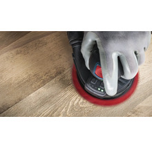 Eine Hand mit Schutzhandschuh bedient einen Exzenterschleifer auf einem Holzboden.