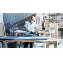 Ein Mann schleift eine blaue Holzplatte mit einem Exzenterschleifer in einer Werkstatt.