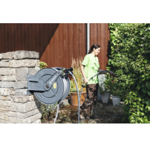 Frau bewässert Pflanzen mit einem Gartenschlauch von einer Schlauchtrommel an einer Steinmauer im Garten. GEKA Logo.