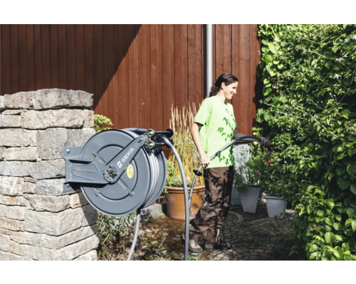 Frau bewässert Pflanzen mit einem Gartenschlauch von einer Schlauchtrommel an einer Steinmauer im Garten. GEKA Logo.