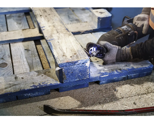 Eine Person sägt eine Holzpalette mit einer Säbelsäge. Ein Brecheisen liegt auf dem Boden.