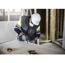 Ein Handwerker schneidet mit einer Stichsäge eine Gipskartonplatte, während er Schutzkleidung trägt.