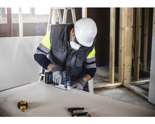 Ein Handwerker schneidet mit einer Stichsäge eine Gipskartonplatte, während er Schutzkleidung trägt.
