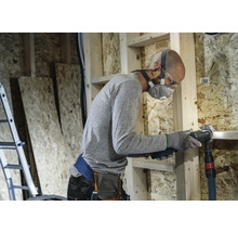 Handwerker mit Atemschutzmaske und Schutzbrille beim Arbeiten mit einem Multifunktionswerkzeug in einem Rohbau