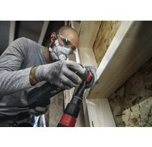 Handwerker bearbeitet Holz mit einem Multifunktionswerkzeug, inklusive Staubabsaugung, Schutzbrille und Atemschutz.