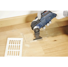 Eine Person schneidet mit einem Multifunktionswerkzeug ein Loch in einen Holzboden neben einem Lüftungsgitter.