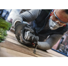 Ein Mann mit Schutzbrille, Atemschutz und Handschuhen arbeitet mit einem Multifunktionswerkzeug an einem Holzbrett.