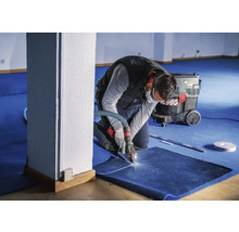 Handwerker schneidet Teppichboden mit einem Spezialwerkzeug, Industriestaubsauger im Hintergrund