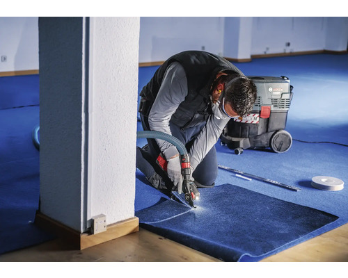 Handwerker schneidet Teppichboden mit einem Spezialwerkzeug, Industriestaubsauger im Hintergrund