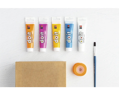 Verschiedene Tuben Farbe, Pinsel, Geschenkband und ein Karton liegen auf einem Tisch.