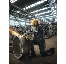 Ein Arbeiter schleift mit einer Schleifmaschine ein Rohr in einer Fabrikhalle, dabei trägt er Schutzkleidung.