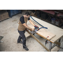 Ein Mann arbeitet mit einer Oberfräse an einem Werkstück auf einer Werkbank in einer Werkstatt.