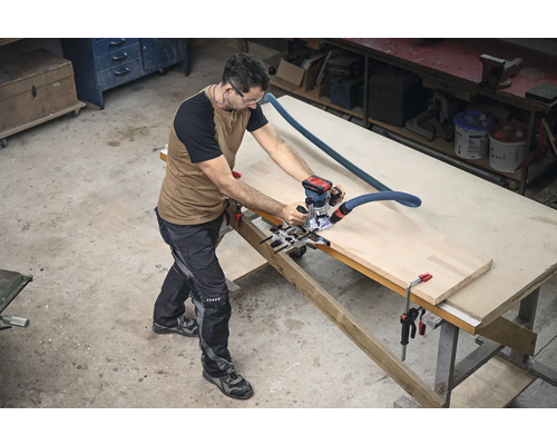 Ein Mann arbeitet mit einer Oberfräse an einem Werkstück auf einer Werkbank in einer Werkstatt.