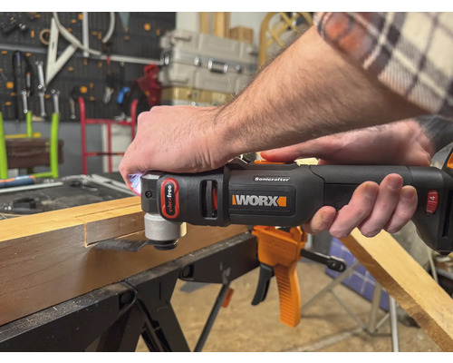 Eine Person benutzt ein Multifunktionswerkzeug der Marke Worx, um ein Stück Holz zu bearbeiten. Das Werkzeug ist schwarz und orange.