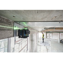 Ein grüner Linienlaser ist an einem Betonbalken befestigt und projiziert eine grüne Laserlinie. Im Hintergrund steht ein Arbeiter auf einem Gerüst.