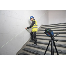 Ein Mann mit Schutzhelm und Warnweste benutzt einen Linienlaser auf einer Treppe, um eine Markierung an einer Wand anzubringen.