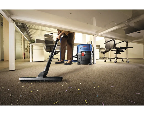 Eine Person reinigt mit einem Staubsauger einen Teppichboden im Büro unter einem Schreibtisch.