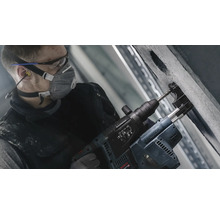 Ein Arbeiter bohrt mit einem Bohrhammer ein Loch in eine Betonwand. Der Arbeiter trägt Schutzkleidung.