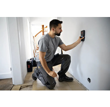 Ein Mann kniet vor einer Wand und verwendet einen Ortungsgerät, um Leitungen zu finden. Eine Werkzeugkiste steht im Hintergrund.