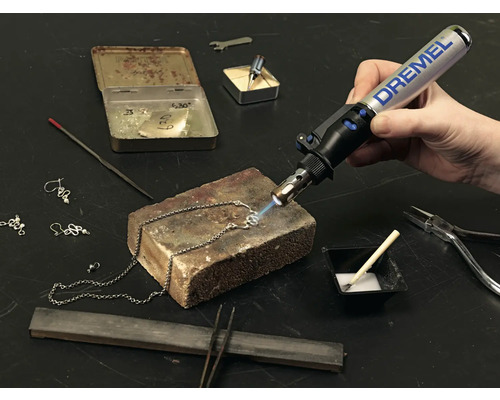 Detaillierte Aufnahme einer Person, die mit einem Dremel-Brenner an einer Halskette arbeitet, zusammen mit Werkzeugen wie Zange und Lötzinn.