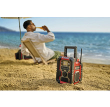 Ein Einhell Baustellenradio am Strand mit einer Person im Hintergrund, die ein Getränk genießt.