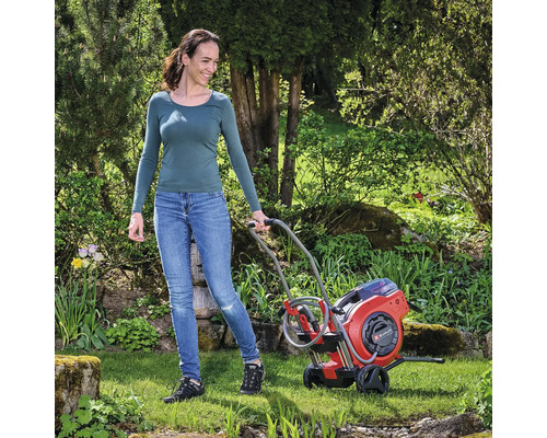 Frau schiebt einen roten Gartenschlauchwagen von Einhell durch den Garten